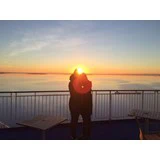 To mennesker står tæt sammen på en terrasse ved solopgang eller solnedgang og vender ud mod et roligt vandområde med solen i horisonten, der skaber en varm glød omkring dem.