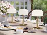 Tre moderne bordlamper med hvide kupler og guldfod står på et udendørs spisebord, der er dækket med tallerkener, kopper, kager og en buket hvide blomster i baggrunden.