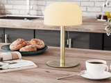 En bordlampe af messing med en rund lampeskærm står på køkkenbordet ved siden af en kop kaffe, wienerbrød på en tallerken og et åbent blad.