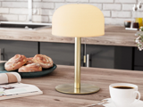 En bordlampe af messing med en hvid lampeskærm står på en køkkenbordplade af træ ved siden af kager, en kop kaffe og et åbent magasin.