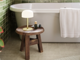 Ved siden af et hvidt badekar, hvor der hænger et håndklæde over kanten, står en træskammel med en lampe og toiletartikler; væggene er beklædt med grønne fliser, og gulvet er belagt med beige fliser.