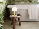 Et moderne badeværelse med et hvidt badekar, grønne flisebelagte vægge, en træskammel med en lampe, et håndklæde og toiletartikler samt et håndklæde, der hænger på badekarret.