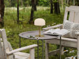 Et rundt bord med en lille lampe, en åben bog og tusindfryd står mellem to træstole med mønstrede puder i et grønt, skovklædt udendørsområde.