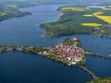 Luftfoto af en lille by på en halvø ved søen, omgivet af vand og grønne marker med gule pletter under en klar himmel.
