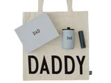 En cremefarvet taske med "DADDY" trykt med store sorte bogstaver, en grå æske med påskriften "DAD", en kop med påskriften "DAD" og et mørkt rør med påskriften "HELLO".
