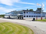 En moderne, hvid kontorbygning i to etager med store vinduer, tre flagstænger og pænt klippet græs under en blå himmel med spredte skyer.