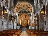 Interiør i en stenkirke med rækker af træbænke, udsmykkede lysekroner, buegange og farverige religiøse vægmalerier over alteret.