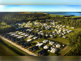 Luftfoto af en stor campingplads med mange autocampere og hytter, omgivet af grønne marker og træer og beliggende tæt på et vandområde.