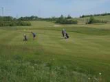 To personer spiller golf på en grøn, græsklædt bane under en klar himmel; den ene putter, mens den anden står i nærheden med en golfvogn.