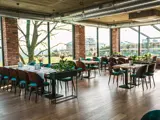 Moderne restaurantinteriør med trægulve, borde og stole, store vinduer, der lukker naturligt lys ind, synlige murstenssøjler og grønne planter som dekoration.