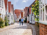 Et par går ned ad en brostensbelagt gade med charmerende huse med røde tegltage og hvide facader på en solrig dag.