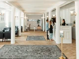 En rummelig hotellobby med hvide søjler, mønstrede tæpper og to personer, der tjekker ind i receptionen sammen med en receptionist.