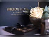 På et skilt står der Chocolate Village by the River Luxury Glamping Resort ved siden af en skål med en hvid rose og en sløret chokoladeæske i forgrunden.