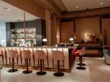 Elegant barinteriør med polstrede taburetter, en velassorteret bar med baggrundsbelysning, varm belysning og et poolbord i baggrunden på et mønstret flisegulv.