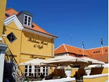 Gult hotel og café med orange tegltag, udendørsservering under store parasoller og klar blå himmel over. På skiltene står der "Folkens Hotel & Cafe" og "Restaurant".
