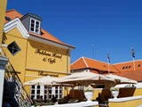 Gul bygning med Foldens Hotel & Café Restaurant på facaden, røde tegltage, store hvide parasoller og mennesker, der spiser udenfor under en klar blå himmel.