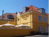 Et lysegult hotel og café med rødt tegltag, udendørs siddepladser under parasoller og cykler parkeret foran på en solrig dag.
