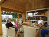 En kvinde, et barn og en mand sidder ved et udendørs træbord på en overdækket terrasse med drinks og magasiner i nærheden af en campingvogn og grønne områder.