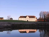 En stor hvid bygning med rødt tag står på en græsklædt bakke og spejler sig i en rolig dam under en klar blå himmel. Bladløse træer ses til højre.