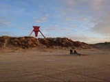 En stor rød stjerneformet skulptur står på en sandklit under en delvis overskyet himmel, og to mennesker sidder i nærheden på stranden.