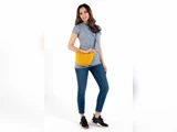 En kvinde i grå t-shirt, blå jeans og sorte sneakers står smilende med en gul crossbody-taske på en hvid baggrund.