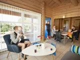 En familie er samlet i en hyggelig træhytte. To voksne og et barn sidder ved et bord, en anden voksen holder et barn i en lænestol, og en kvinde står ved døren. Sollyset strømmer ind gennem store vind