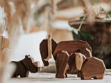 Tre dyrefigurer af træ, bl.a. en elefant og en flodhest, er anbragt på en blød overflade med slørede planter i baggrunden.