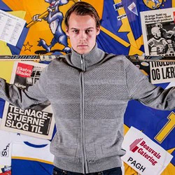 En mand i en grå sweater holder en hockeystav over skuldrene og står foran en farverig baggrund med plakater, avisudklip og tegneseriegrafik.