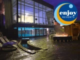 Udendørs spa-område om aftenen med et rundt boblebad, liggestole og en moderne glasbygning; Enjoy Resorts logo i øverste højre hjørne.
