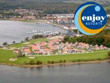 Luftfoto af et kystresort med huse med røde tage nær vandet, træer og havne, med Enjoy Resorts logo i øverste højre hjørne.