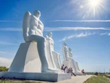 Fire store hvide statuer af siddende mænd under en lyseblå himmel, med mennesker siddende ved deres fødder og solen skinnende over dem.
