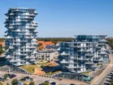 To moderne, kantede boligblokke med karakteristiske geometriske altaner, omgivet af grønne områder, parkerede biler og mindre bygninger under en klar blå himmel.