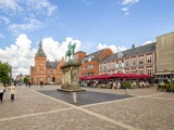 Et brostensbelagt torv med en rytterstatue i midten, omgivet af røde murstensbygninger, udendørs caféer og mennesker, der går rundt under en delvis overskyet himmel.