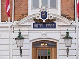 Indgangen til Hotel Eyde med et blåt og gyldent skilt over en trædør, flankeret af to lamper og med røde og hvide flag på hver side.