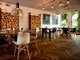 Moderne restaurant med trægulve, runde borde, blandede sorte og hvide stole, vægdekoration i træ, pendler og store vinduer, der lukker naturligt lys ind.