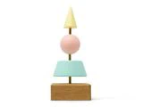 En minimalistisk skulptur med en træbase, blå trapez, pink kugle og gul kegle stablet lodret på en tynd stang.