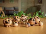 En gruppe små, runde dyrefigurer af træ med store øjne står på et træbord med grønne planter og et sløret rum i baggrunden.