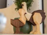 To fuglefigurer i træ, den ene lys og den anden mørkere, står side om side foran en grøn vase med en bladplante.