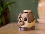 En lille, rund hundefigur af træ med mørke ører, store øjne og en flad træhue, der sidder på en træplade.