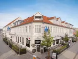 En hvid hjørnebygning med røde tagsten huser et Best Western-hotel og en kaffebar med udendørs siddepladser og træer langs den brostensbelagte gade.