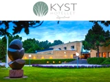 Moderne hotelbygning i mursten med store vinduer, omgivet af grøn plæne og træer, med en rund abstrakt skulptur i forgrunden. Logo Kyst Hotellet Djursland er øverst.