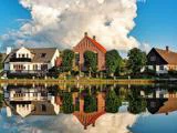 Charmerende huse og træer spejler sig i en rolig sø med en dramatisk, stor sky, der fylder den blå himmel ovenover.