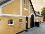 En lang, gul stukbygning med hvide kanter, sorte buede døre og små vinduer. Nummeret 1926 står over den store dør. En potteplante står nær indgangen.