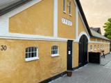 En lang gul bygning med hvid kant, sort buet dør og små vinduer. Nummeret 1924 står over døren, og nummer 30 står på venstre væg.