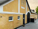 En lang gul bygning med hvid kant, sort buet dør og små vinduer. Nummeret 1924 står over døren, og nummer 30 står på venstre væg.