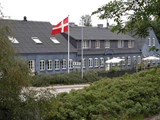 Et dansk flag vajer foran en blågrå bygning med buede vinduer og et KRO-skilt, omgivet af grønne områder og udendørs siddepladser med hvide parasoller.