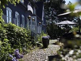 En charmerende brostensbelagt gårdhave ved siden af en blå bygning med buede vinduer, lilla blomster i potter og udendørs siddepladser i skyggen af parasoller.