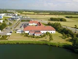En stor bygning med rødt tegltag ligger ved en dam, omgivet af græs, træer, parkerede biler, veje og åbne marker under en delvis overskyet himmel.