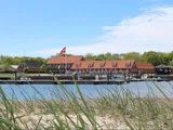 En sandstrand med græs i forgrunden, en bådbro med både, røde bygninger, træer og et dansk flag i baggrunden under en delvis overskyet himmel.