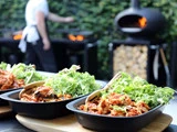Tre sorte fade fyldt med bladgrøn salat og strimlet mad står på et træbræt. I baggrunden laver en person mad i nærheden af en brændefyret ovn og grill udendørs.